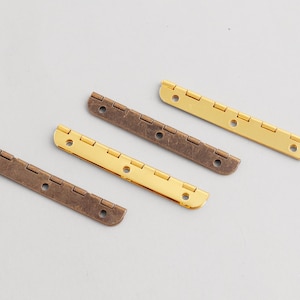 May include: Four metal hinges in two colors, bronze and gold, are arranged diagonally on a light gray surface. Each hinge has a series of small holes for screws and a folding design. The hinges are for woodworking or craft projects.