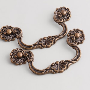 May include: Two antique bronze-colored metal drawer pulls featuring a curved handle design with ornate floral and shell-like detailing on the mounting plates and the center of the handle. The hardware has a dark, weathered finish with highlights.