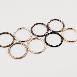 Könnte beinhalten: Eine Auswahl an Metall-O-Ringen in verschiedenen Farben, darunter Bronze, Silber, Dunkelgrau, Schwarz und Gold. Diese kreisförmigen Ringe sind auf einer weißen Oberfläche angeordnet und zeigen ihre unterschiedlichen Oberflächen und potenziellen Anwendungen.