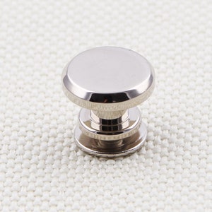 May include: A shiny, silver-colored metal button stud with a rounded top and a tiered base. The button is designed for use in clothing or accessories, offering a sleek and modern aesthetic.