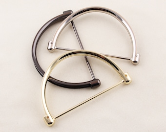 May include: Three metal purse handles in black, silver, and gold. Each handle is a half-circle shape with a straight bar connecting the ends. The handles are arranged in a layered pattern against a neutral background.