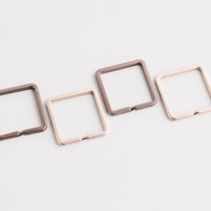 May include: Four square metal rings in shades of brown and gold are arranged diagonally on a white surface. Each ring has a small opening on one side. The rings are likely used for crafting or as hardware.