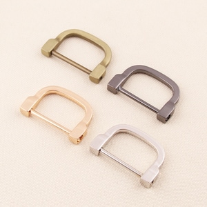 May include: Four metal D-ring buckles in different finishes: antique brass, rose gold, gunmetal, and silver. Each buckle has a rectangular shape with a rounded top and a straight bar. These buckles are suitable for bags, belts, or leathercraft.