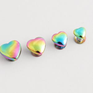 May include: Four heart-shaped cabinet knobs with a vibrant, iridescent finish. The knobs transition through a spectrum of colors, including pink, yellow, green, and blue. They are mounted on a silver base, perfect for adding a touch of color.