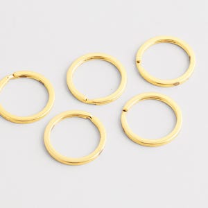 May include: Five gold-colored key rings are arranged on a light gray surface. Each ring is circular, with a hinged opening for attaching keys. The rings have a shiny, metallic finish, reflecting the light.