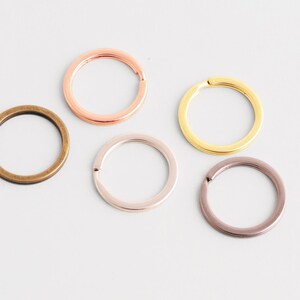 May include: Five metal key rings in various colors: bronze, rose gold, gold, silver, and a dark purple. The rings are round with a small opening for attaching keys or other items. The key rings are on a white surface.