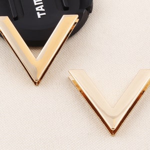 May include: Two gold-colored metal V-shaped embellishments. The pieces have a shiny, reflective surface and are set against a neutral background. These decorative elements could be used for crafting or fashion accessories.