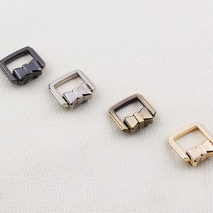 May include: Four metal bow-shaped buckles in different finishes: black, silver, gold, and rose gold. The buckles are square-shaped with a bow design in the centre.