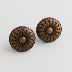Könnte beinhalten: Zwei runde Schrankknöpfe in Bronzefarbe. Jeder Knopf hat ein erhabenes Blumendesign mit einem zentralen Kreis und blütenblattartigen Formen, die nach außen strahlen. Die Knöpfe haben ein Vintage- und antikes Aussehen.