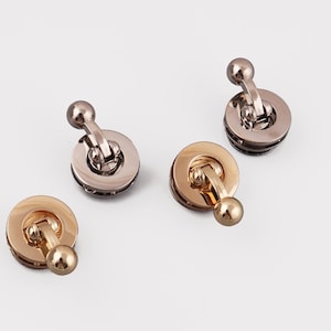 Pode incluir: Quatro fechos de metal para bolsas em tons de prata e dourado. Cada fecho apresenta uma base circular com um braço articulado e um botão esférico. Os fechos estão dispostos sobre um fundo branco, mostrando o seu design e acabamento.