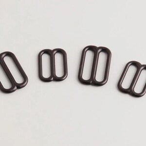 May include: Four dark brown metal strap slides, each with two rectangular openings. The slides are arranged on a light background. These hardware pieces are likely used in bag making or garment construction.