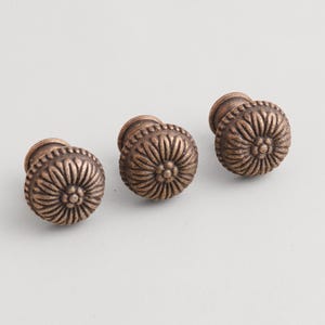 May include: Three bronze-colored cabinet knobs with a floral design. Each knob has a rounded, textured surface with a central flower-like pattern and a short, cylindrical base. The knobs are arranged on a light background.