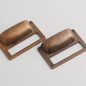 Peut inclure: Deux poignées de tiroir antiques de couleur bronze. Chaque poignée a un cadre rectangulaire avec une poignée encastrée et est fixée avec de petites vis. La quincaillerie a un aspect vintage et vieilli, adapté à la restauration de meubles.