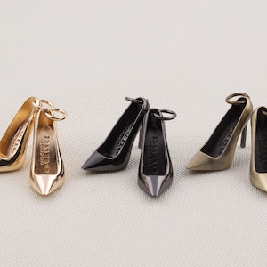 May include: A collection of miniature high heel shoe charms in gold, black, and bronze tones. Each charm features a pointed toe and a thin heel, with the text "Dune London" visible on the sole. The charms are designed with a small loop for attachment.