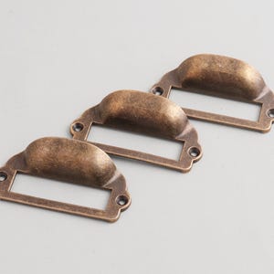 May include: Three antique bronze-colored drawer pulls with label holders. Each pull has a curved handle and a rectangular space for a label. The hardware is made of metal and has a vintage aesthetic.