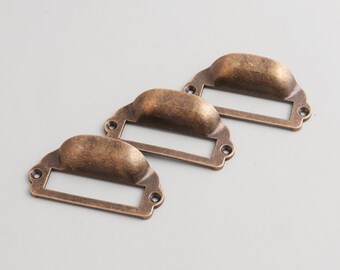 May include: Three antique bronze-colored drawer pulls with label holders. Each pull has a curved handle and a rectangular space for a label. The hardware is made of metal and has a vintage aesthetic.