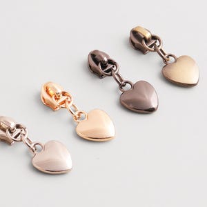 Könnte beinhalten: Vier Metall-Reißverschlussanhänger mit herzförmigen Anhängern in verschiedenen Metallic-Ausführungen: Roségold, Kupfer, Bronze und Gold. Ideal zum Personalisieren von Kleidungsstücken, Taschen oder Accessoires.
