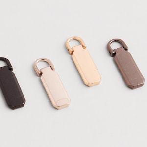 Può includere: Quattro tiranti per cerniera in metallo di vari colori: marrone scuro, oro chiaro, oro rosa e bronzo. Ogni tirante ha un corpo rettangolare con una parte superiore arrotondata e un piccolo anello.