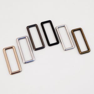 May include: Six rectangular metal rings in various finishes: rose gold, silver, black, and bronze. These hardware components are used in bag making and other crafting projects. The rings are arranged on a white surface.