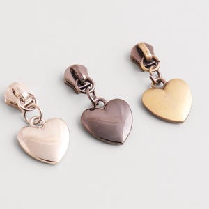 Può includere: Tre tiretti per cerniera in metallo con ciondoli a forma di cuore. I tiretti sono in argento, canna di fucile e oro. I ciondoli a forma di cuore sono lisci e lucidi.