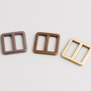 May include: Three metal rectangle sliders in bronze, copper, and gold tones. Each slider has two rectangular openings. These hardware pieces are likely used in bag making or other crafting projects.