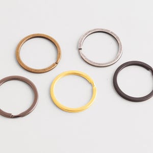 Puede incluir: Cinco llaveros metálicos en varios colores: bronce, plata, marrón oscuro, dorado y negro. Cada anillo es circular con una abertura para sujetar llaves. Los anillos están dispuestos sobre una superficie blanca.