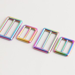 May include: Four iridescent metal buckles in a rainbow colour scheme. The rectangular buckles have a sliding bar and are in different sizes. The buckles are set against a plain white background.