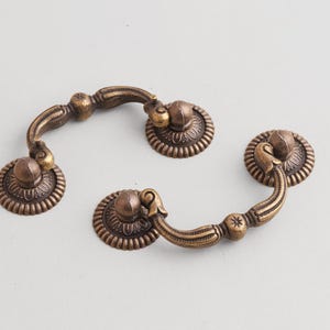 Pode incluir: Três puxadores de gaveta antigos em bronze. Cada puxador apresenta uma pega curva e uma placa traseira circular decorativa com um padrão detalhado. O hardware tem uma estética vintage, adequado para restauração de móveis ou novas construções.