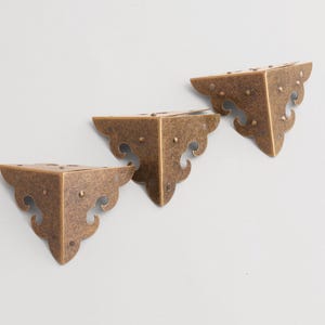 May include: Three antique bronze corner brackets. Each bracket is triangular with decorative scrollwork along the edges and small rivet details. The brackets are arranged diagonally against a light background.