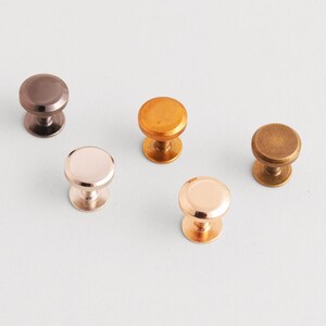 Könnte beinhalten: Eine Auswahl von fünf Metall-Schrankknöpfen in verschiedenen Ausführungen: Bronze, Kupfer, Roségold und Gold. Jeder Knopf hat eine abgerundete Oberseite und einen zylindrischen Sockel, geeignet für Möbel oder Dekoration.