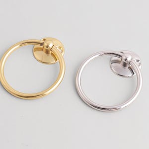 Può includere: Due pomelli per mobili a forma di anello. Uno è dorato, l'altro argentato. Ogni pomello ha un anello circolare attaccato a una base circolare con un piccolo pomello arrotondato. I pomelli sono su uno sfondo grigio chiaro.