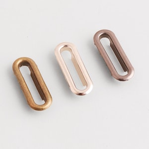 May include: Three metal oval eyelets in bronze, rose gold, and copper colors. These hardware pieces have a rectangular opening in the center. The eyelets are used for various crafting and sewing projects.
