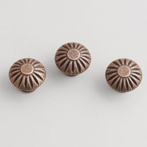 May include: Three bronze-colored cabinet knobs with a sunburst design. Each knob is round with a central circular detail and radiating lines. The knobs are made of metal and have a vintage or antique appearance.