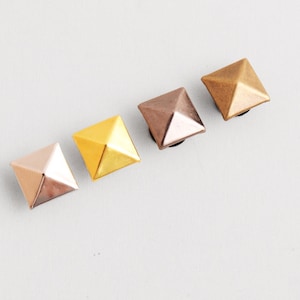 Può includere: Quattro borchie metalliche a forma di piramide in fila. Le borchie sono nelle tonalità oro rosa, oro, bronzo e rame. Le borchie sono su una superficie bianca.