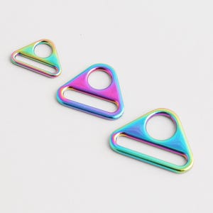 May include: Three iridescent rainbow-colored metal triangle rings with a circular hole and a rectangular opening. The hardware is displayed on a white background. These rings are suitable for various crafting and accessory projects.