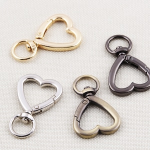 Könnte beinhalten: Vier herzförmige Drehverschlüsse in Gold, Silber, Bronze und Gunmetal-Farben. Jeder Verschluss hat einen Ring zur Befestigung an einem Schlüsselanhänger oder anderen Gegenständen. Die Verschlüsse sind aus Metall und werden auf einem neutralen Stoffhintergrund gezeigt.