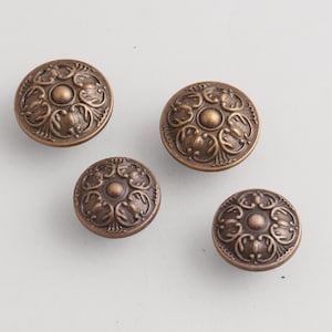 May include: Four antique bronze cabinet knobs with ornate floral designs. Each knob is round with a raised central circle and detailed patterns. The knobs are made of metal and have a warm, aged finish.