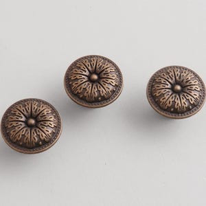 May include: Three bronze cabinet knobs with a floral design. Each round knob features a detailed pattern radiating from the centre. The knobs are set against a neutral background.