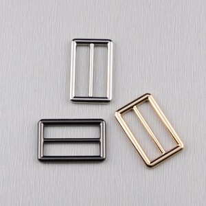 May include: Three metal rectangle slider buckles in silver, black, and gold. Each buckle has a single bar in the centre and is designed for strap adjustment. The buckles are displayed on a textured grey surface.