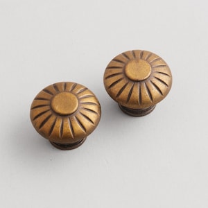 May include: Two antique-style cabinet knobs with a bronze finish. Each knob features a circular design with a central disc and radiating lines, resembling a stylized flower. The knobs are likely made of metal and are suitable for furniture restoration.