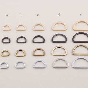 Puede incluir: Surtido de anillas en D en varios acabados metálicos, incluyendo dorado, bronce, plateado y negro. Las anillas en D están dispuestas en filas y columnas, mostrando diferentes tamaños y colores. Estas piezas de ferretería metálica se utilizan comúnmente en la fabricación de bolsos y manualidades.