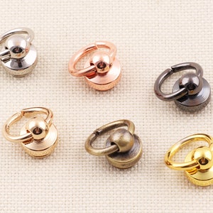 Könnte beinhalten: Eine Auswahl an Metall-Drehverschlüssen in verschiedenen Ausführungen: Silber, Roségold, Gunmetal, Gold und Antik-Messing. Jeder Verschluss hat eine runde Basis mit einem Ring und einer kugelförmigen Oberseite.