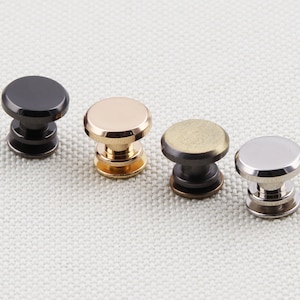 May include: Four metal rivets in a row, each with a different finish: black, gold, bronze, and silver. The rivets have a rounded top and a cylindrical base, suitable for leathercraft or other DIY projects.