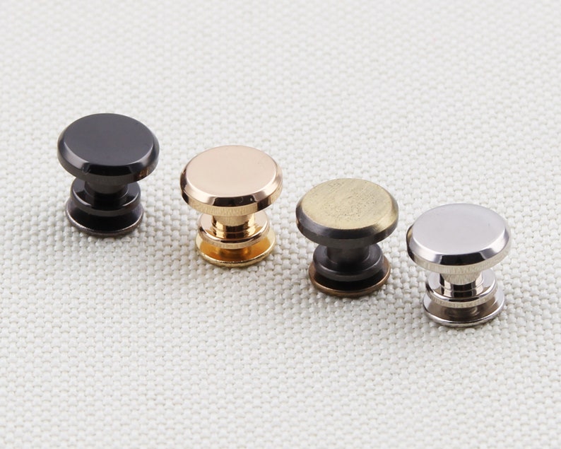May include: Four metal rivets in a row, each with a different finish: black, gold, bronze, and silver. The rivets have a rounded top and a cylindrical base, suitable for leathercraft or other DIY projects.
