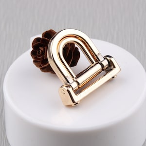 Puede incluir: Una hebilla en forma de D de metal dorado, con una base rectangular y una parte superior curvada. La hebilla está diseñada para su uso en bolsos, cinturones u otros accesorios. Una pequeña piña marrón descansa detrás de la hebilla.