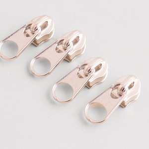 May include: Four rose gold zipper pulls arranged diagonally on a light gray surface. Each pull has a rounded oval shape with a circular opening and a rectangular base.