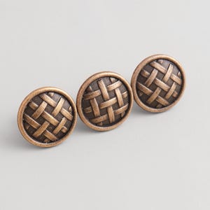 Peut inclure: Trois boutons de meuble en bronze avec un motif de panier tressé. Les boutons ronds présentent un motif texturé. La quincaillerie a une esthétique vintage, adaptée à la restauration de meubles ou aux nouvelles constructions.