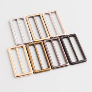 Puede incluir: Colección de hebillas correderas rectangulares de metal en varios acabados metálicos, incluyendo plata, oro, bronce y negro. Estos componentes de hardware presentan un diseño de tres barras, adecuados para correas o cinturones.