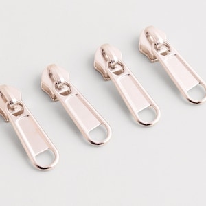 May include: Four metallic rose gold zipper pulls arranged on a light gray surface. Each pull has a rectangular body with a rounded loop at the end and a circular top. The pulls are evenly spaced and appear new.