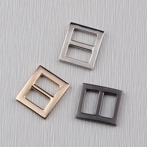 May include: Three metal rectangular buckles in silver, gold, and gunmetal grey. Each buckle has two rectangular openings. The buckles are arranged on a textured, light grey surface.